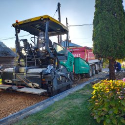 Firma Handlowo-Produkcyjno-Uslugowa 'Kocjan' - Rozściełacz asfaltu Vogele Super 1600-3 w trakcie pracy, obok wywrotka KP-KIPPER PF67, widoczny pracownik w odblaskowej kamizelce, w tle budynek gospodarczy i niebieskie niebo.