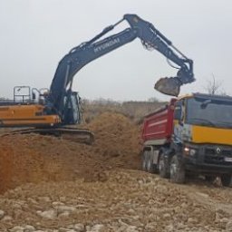 Firma Handlowo-Produkcyjno-Uslugowa 'Kocjan' - Żółto-czarna koparka Hyundai ładuje urobek do czerwonej wywrotki Renault na nierównym, piaszczystym terenie w pochmurny dzień.