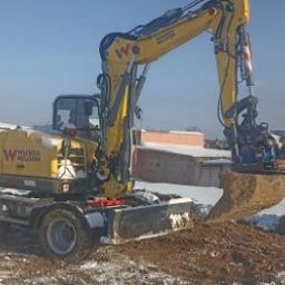 Firma Handlowo-Produkcyjno-Uslugowa 'Kocjan' - Żółta koparka kołowa Wacker Neuson z łyżką nabiera ziemię na zasnieżonym placu budowy w słoneczny dzień. W tle widoczne budynki mieszkalne.