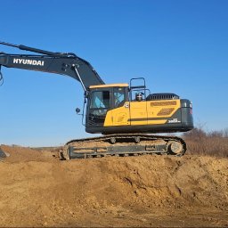 Firma Handlowo-Produkcyjno-Uslugowa 'Kocjan' - Żółto-czarna koparka Hyundai HX300A na wzniesieniu z ziemi, z łyżką skierowaną w dół, na tle błękitnego nieba i zabudowań w oddali.