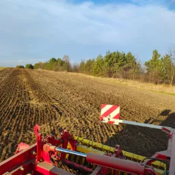 Widok z kabiny ciągnika na pole po niwelacji, z widocznymi rzędami świeżo obrobionej ziemi i lasem w tle, oświetlonym popołudniowym słońcem.