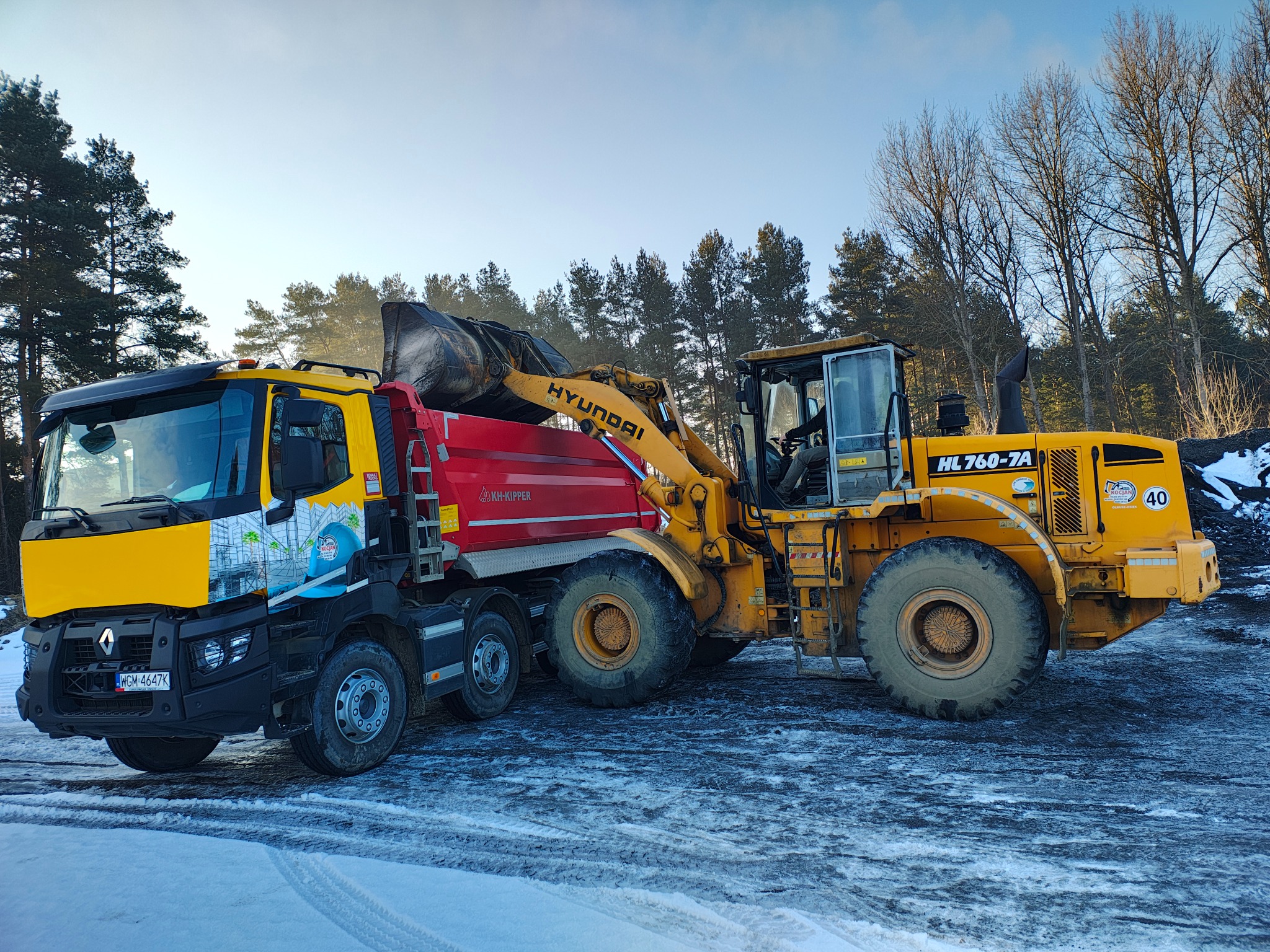 Żółta ładowarka Hyundai HL760-7A ładuje łyżką materiał do czerwonej wywrotki Renault w zimowy dzień, widoczne drzewa w tle.