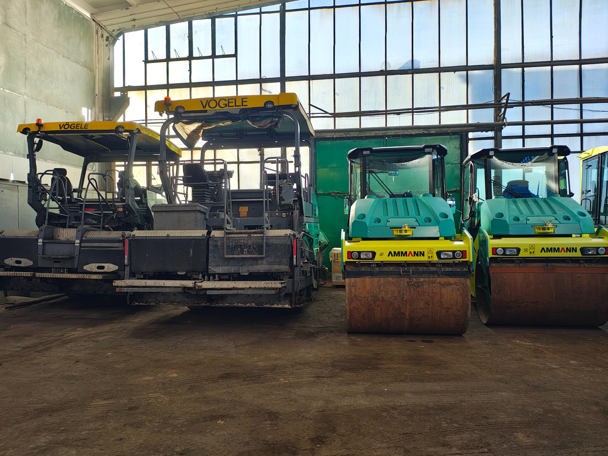 Wnętrze hali z zaparkowanym asfaltobetonowym rozściełaczem marki Vögele oraz dwoma walcami drogowymi marki Ammann, ustawionymi obok siebie.