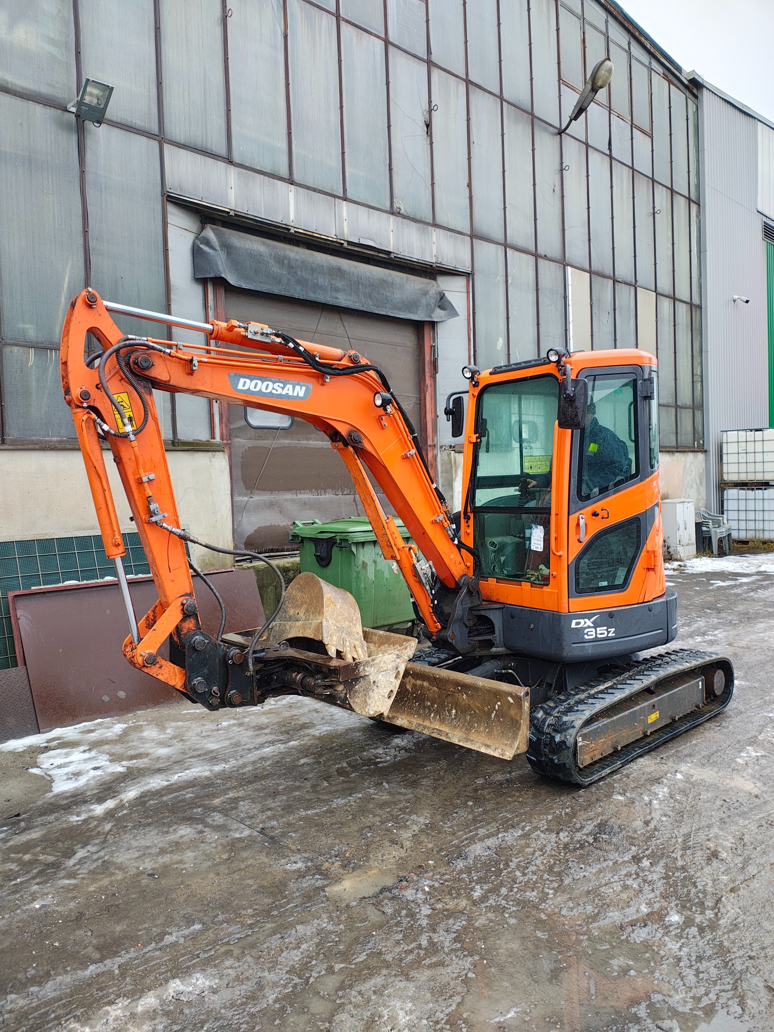 Pomarańczowa mini koparka Doosan DX35Z na tle hali przemysłowej z widocznymi śladami błota i śniegu na ziemi.