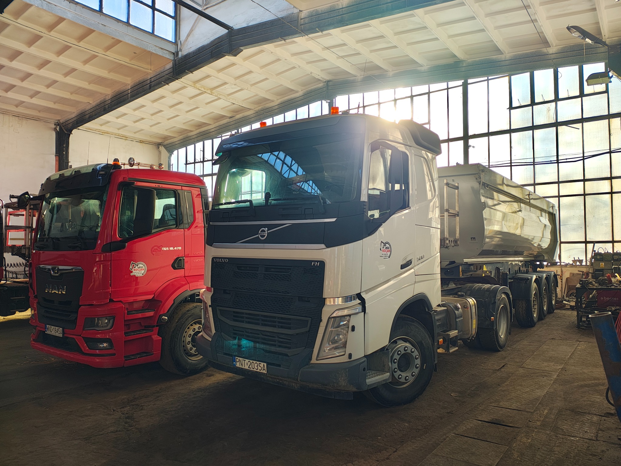 Dwa samochody ciężarowe w hali: czerwony MAN TGS 18.470 z naklejką 'Kocjan' i biały Volvo FH 500, stojące obok siebie, w tle okna z widocznym światłem słonecznym.