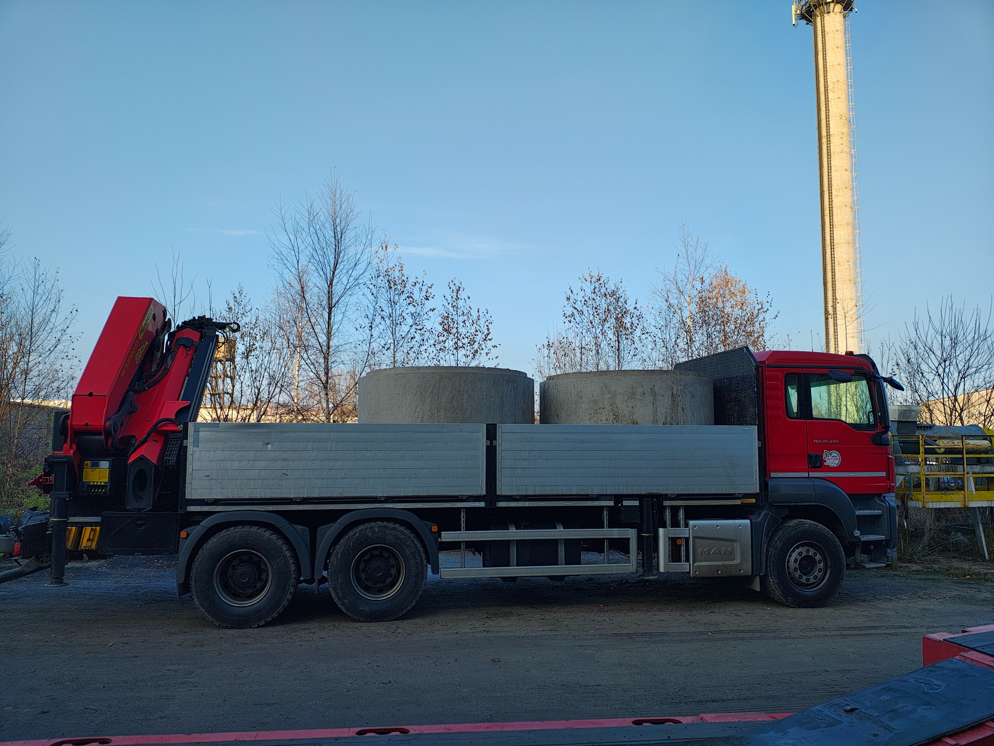 Czerwony samochód ciężarowy MAN z dźwigiem i dwoma betonowymi kręgami w skrzyni ładunkowej, na tle drzew i wysokiego komina.