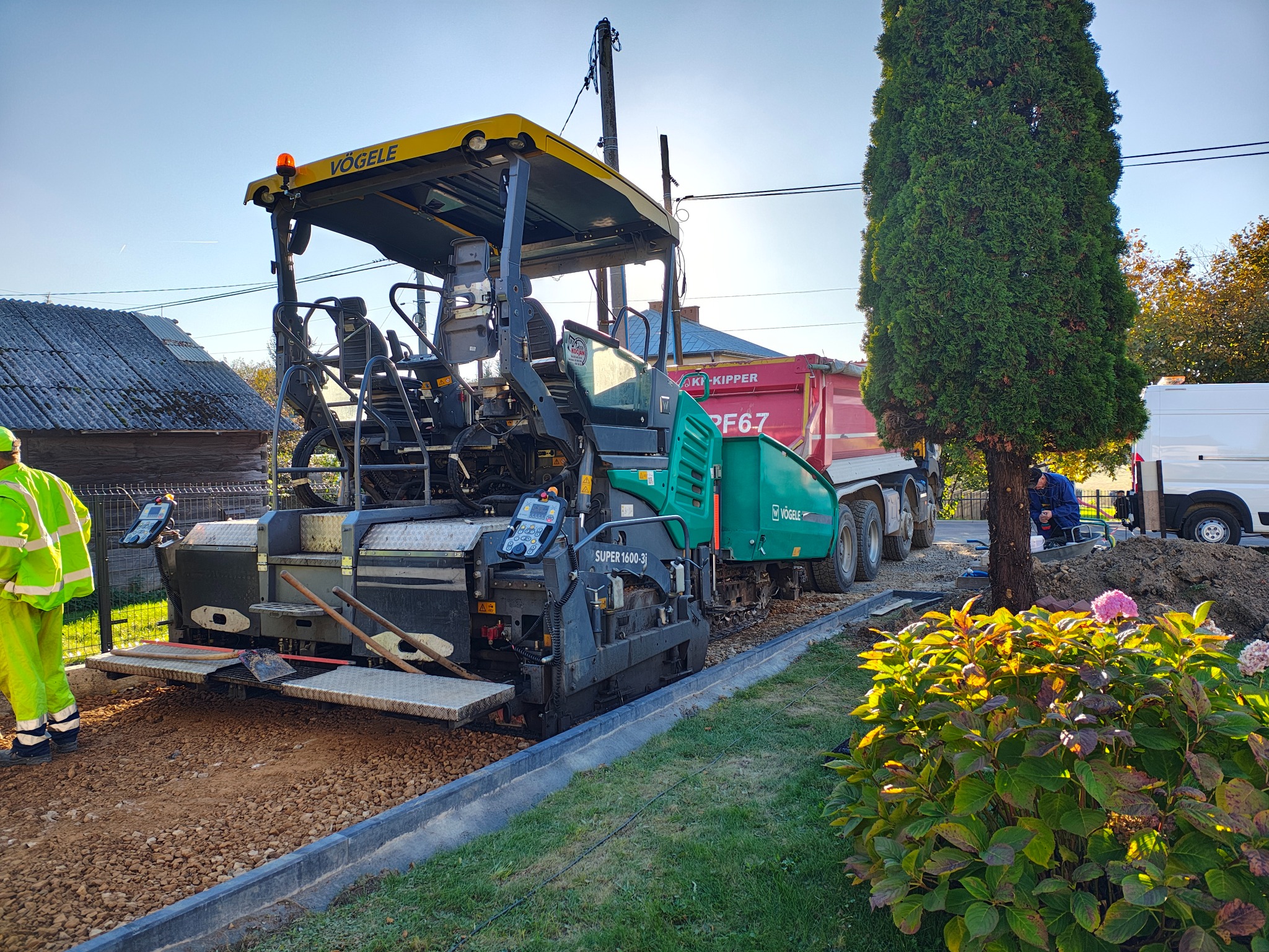 Rozściełacz asfaltu Vogele Super 1600-3 w trakcie pracy, obok wywrotka KP-KIPPER PF67, widoczny pracownik w odblaskowej kamizelce, w tle budynek gospodarczy i niebieskie niebo.