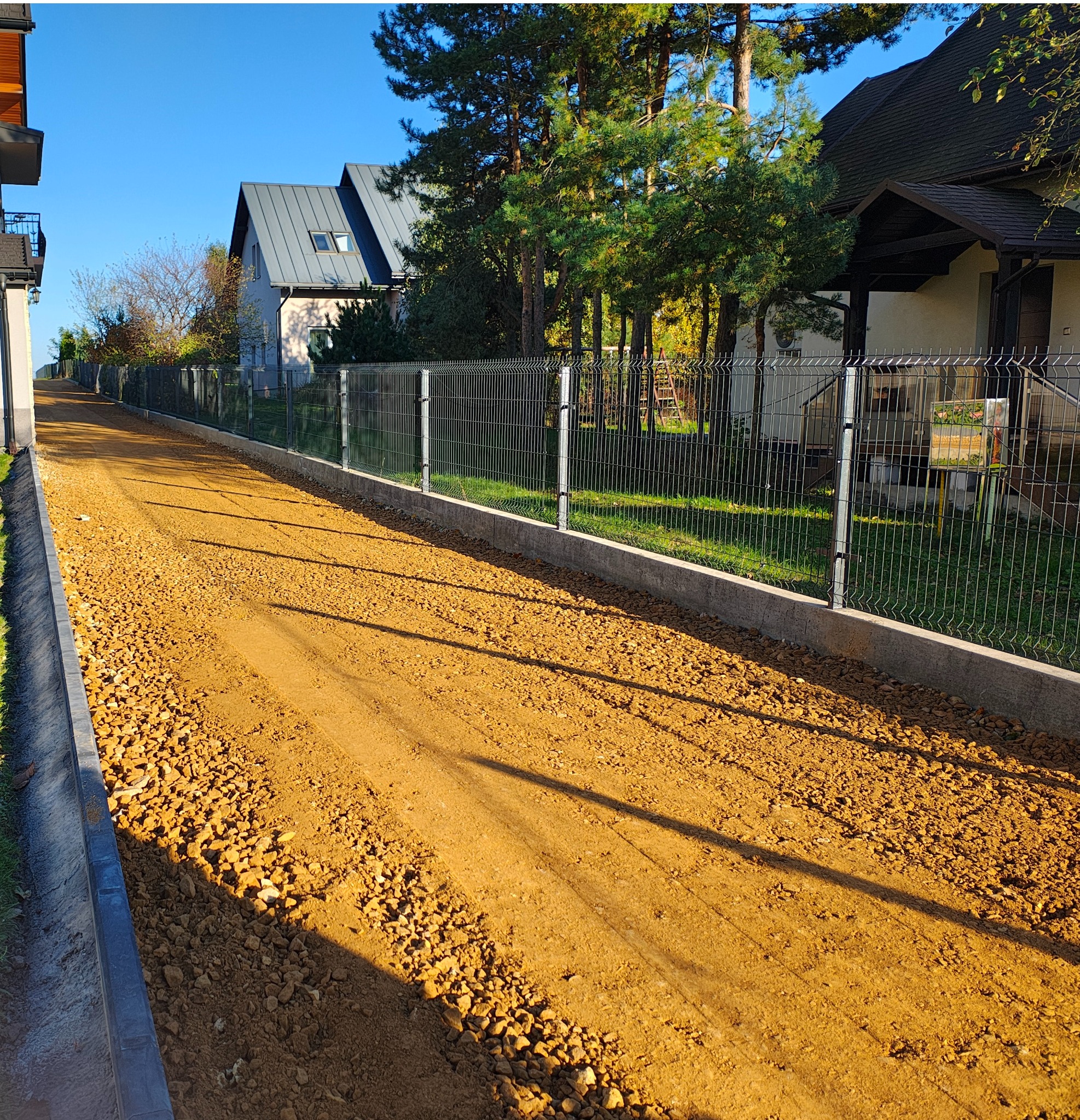 Żwirowa droga dojazdowa, ograniczona krawężnikami i ogrodzeniem panelowym, w tle zabudowa jednorodzinna w słoneczny dzień.