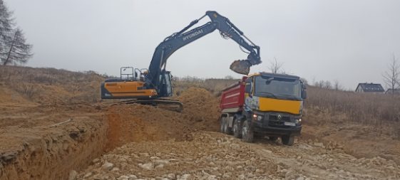 Żółto-czarna koparka Hyundai ładuje urobek do czerwonej wywrotki Renault na nierównym, piaszczystym terenie w pochmurny dzień.