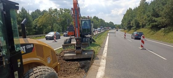 Koparka kołowa i walec CAT CS64B pracują przy krawędzi jezdni, w tle ruch samochodowy i drzewa.