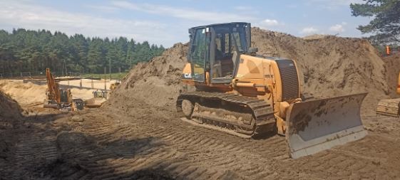 Pomarańczowy spychacz gąsienicowy CASE na placu budowy, z wykopem w tle i lasem na horyzoncie, widoczne ślady gąsienic na ziemi.