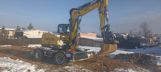 Żółta koparka kołowa Wacker Neuson z łyżką nabiera ziemię na zasnieżonym placu budowy w słoneczny dzień. W tle widoczne budynki mieszkalne.