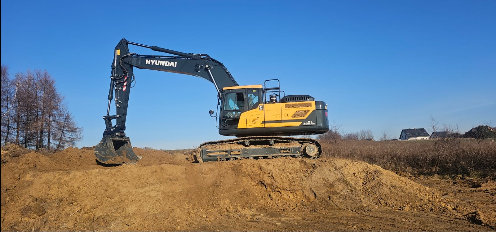 Żółto-czarna koparka Hyundai HX300A na wzniesieniu z ziemi, z łyżką skierowaną w dół, na tle błękitnego nieba i zabudowań w oddali.