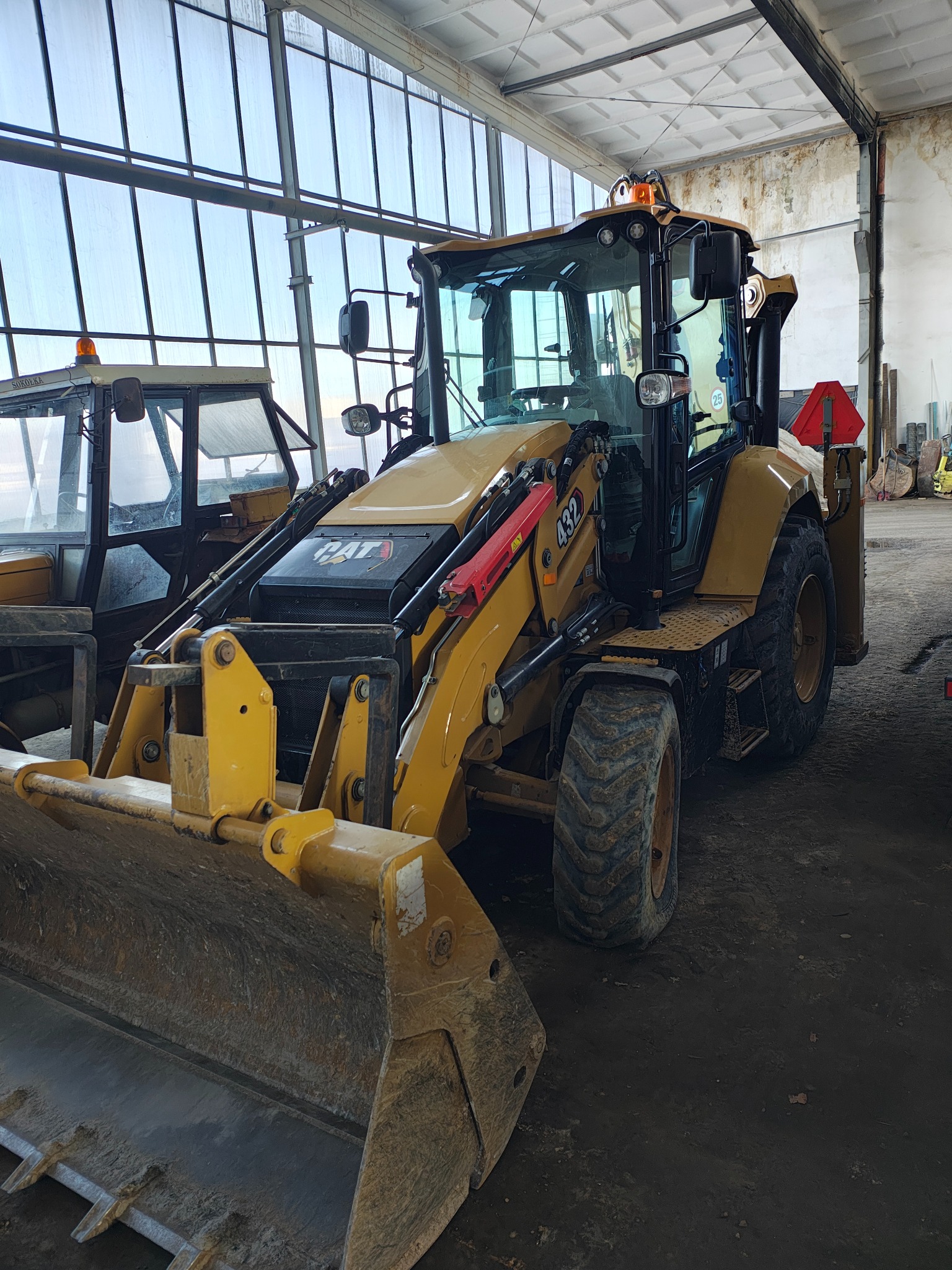 Żółta koparko-ładowarka CAT 432 stoi w hali z dużą ilością okien, widoczny brudny osprzęt i opony, obok zaparkowana inna maszyna budowlana.