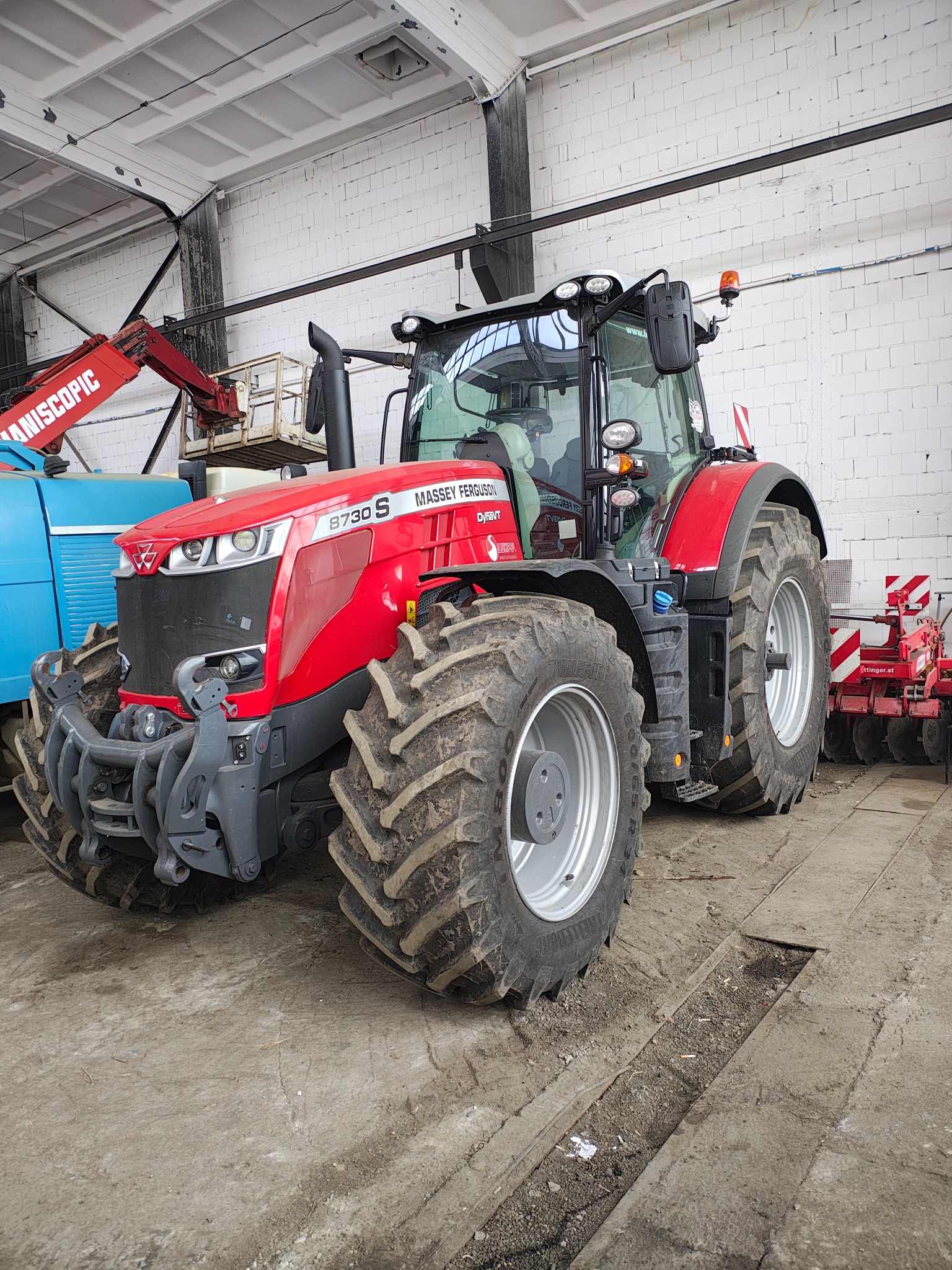 Czerwony traktor Massey Ferguson 8730 S z brudnymi oponami stoi w hali, w tle widoczny podnośnik koszowy i maszyna rolnicza.