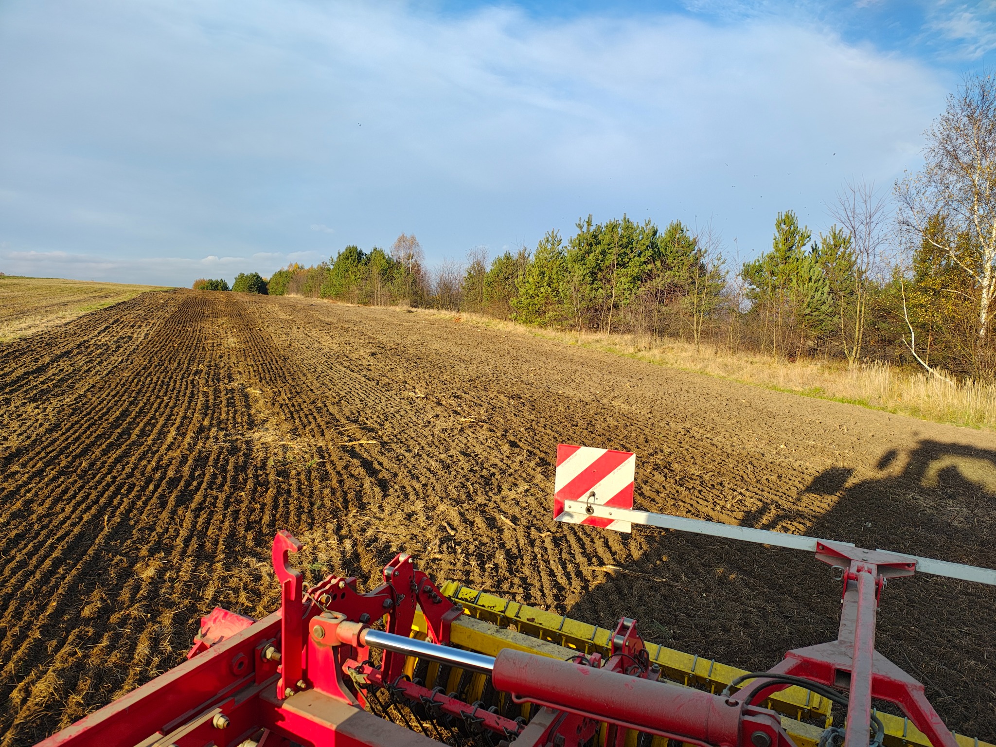 Widok z kabiny ciągnika na pole po niwelacji, z widocznymi rzędami świeżo obrobionej ziemi i lasem w tle, oświetlonym popołudniowym słońcem.