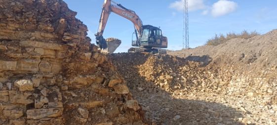 Pomarańczowa koparka CASE na szczycie wykopu, nabiera urobek z ziemi, widoczna warstwowa struktura wykopu i błękitne niebo.