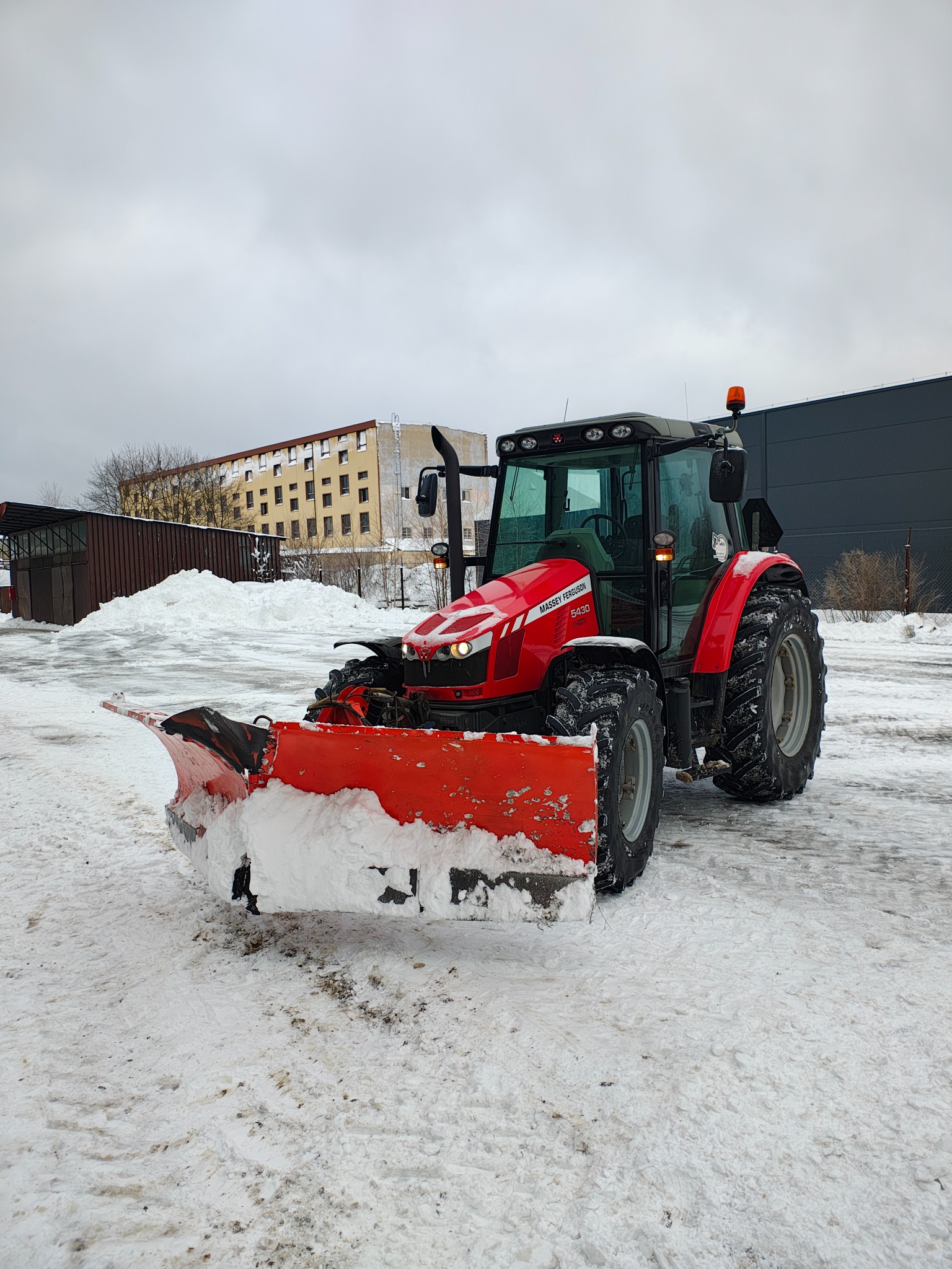 Czerwony traktor Massey Ferguson 5430 z pługiem odśnieżnym stoi na ośnieżonym placu, w tle budynki i pochmurne niebo.