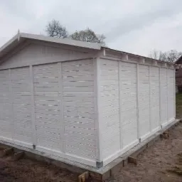 Nowo pomalowany na biało drewniany budynek gospodarczy z deskami ułożonymi poziomo, posadowiony na betonowej wylewce, widok z boku na tle domu jednorodzinnego.