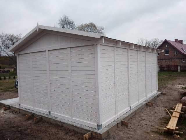 Nowo pomalowany na biało drewniany budynek gospodarczy z deskami ułożonymi poziomo, posadowiony na betonowej wylewce, widok z boku na tle domu jednorodzinnego.