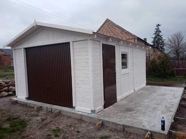 Nowo wybudowany, biały garaż z brązową bramą i drzwiami, betonowa wylewka przed wejściem, widok na budynek mieszkalny w tle.