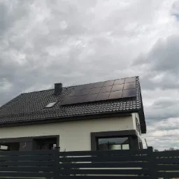 Instalacja fotowoltaiczna typu Full Black na dachu domu jednorodzinnego z ciemną dachówką i oknem dachowym, widok zza szarego ogrodzenia w pochmurny dzień.