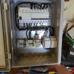 Otwarta skrzynka elektryczna z zamontowanymi bezpiecznikami, stycznikami i przewodami, widoczne narzędzia na drewnianym blacie, w tym kombinerki Gedore z niebieskimi rękojeściami i śrubokręt...