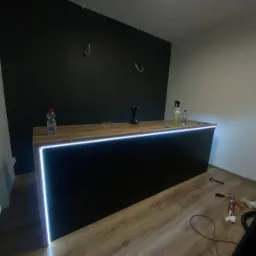 Nowoczesna lada z podświetleniem LED, czarny front, drewniany blat, widoczne narzędzia i materiały montażowe na podłodze, surowa ściana w tle z hakami, oświetlenie sufitowe.