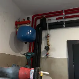 Instalacja grzewcza w kotłowni: czerwone i czarne rury, niebieski zbiornik wyrównawczy, manometr, oraz narzędzia - wiertarka i poziomica laserowa na szczycie bojlera.