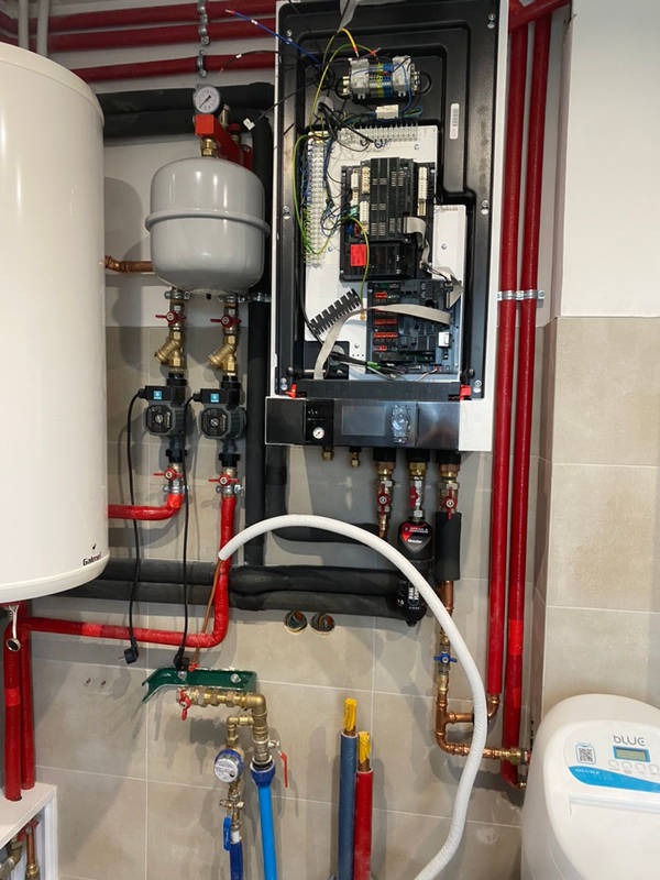 Wnętrze kotłowni z otwartym panelem sterowania kotła gazowego, widoczne liczne rury miedziane i plastikowe, zbiornik wyrównawczy oraz zmiękczacz wody Blue.