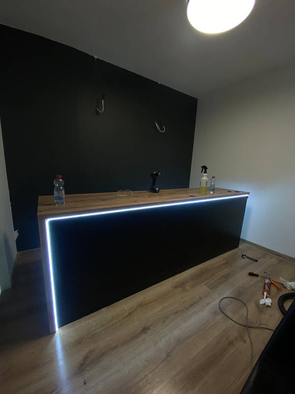 Nowoczesna lada z podświetleniem LED, czarny front, drewniany blat, widoczne narzędzia i materiały montażowe na podłodze, surowa ściana w tle z hakami, oświetlenie sufitowe.