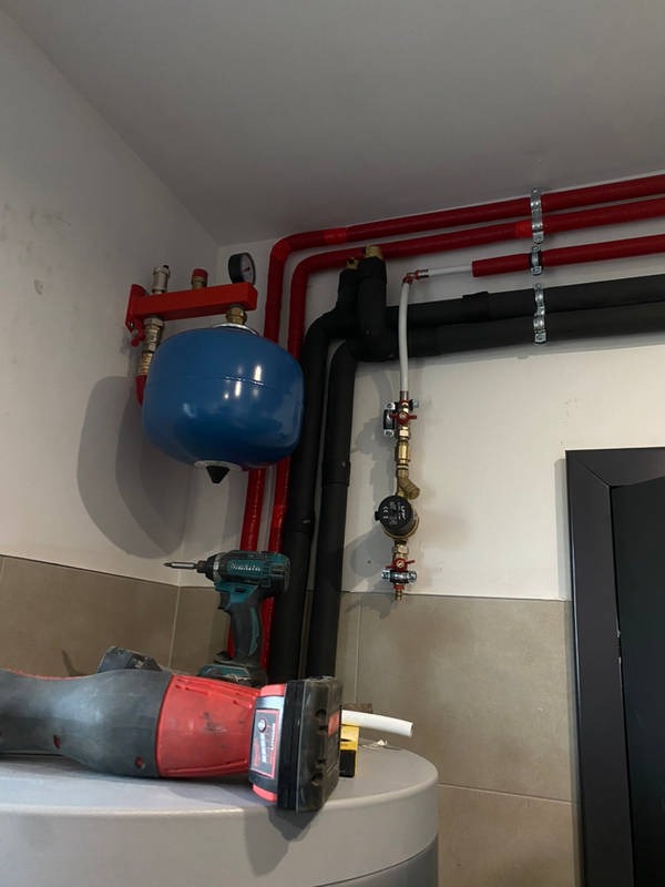Instalacja grzewcza w kotłowni: czerwone i czarne rury, niebieski zbiornik wyrównawczy, manometr, oraz narzędzia - wiertarka i poziomica laserowa na szczycie bojlera.