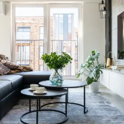 Jasne wnętrze salonu z czarną skórzaną sofą, stolikami kawowymi z filiżankami, kwiatami w wazonie i telewizorem z odbijającym się widokiem budynku na ekranie.