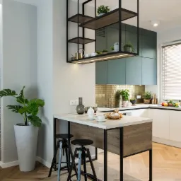 Nowoczesna kuchnia z wyspą, zielonymi szafkami i złotą mozaiką na ścianie, uzupełniona o czarne metalowe półki wiszące i dekoracyjną tapetę z motywami roślinnymi, oświetlona ciepłym światłem.