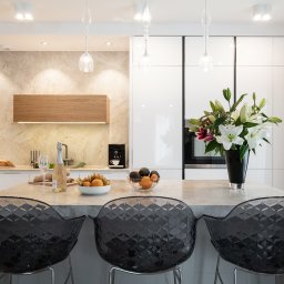 Kodo - Nowoczesna kuchnia z wyspą, marmurowym blatem, transparentnymi krzesłami barowymi i bukietem lilii w czarnym wazonie, oświetlona wiszącymi lampami.