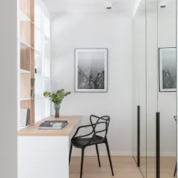 Wąskie, minimalistyczne biuro z drewnianym blatem biurka, czarnym krzesłem o ażurowym oparciu, regałem z jasnego drewna i lustrzanymi drzwiami szafy wnękowej, doświetlone punktowymi lampami...