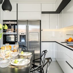 Kodo - Nowoczesna, biała kuchnia z czarnym stołem jadalnym, na którym stoją kwiaty, szklanki i talerze. Widoczny srebrny sprzęt AGD i oświetlenie LED pod szafkami. W prawym górnym rogu logo firmy KODO.