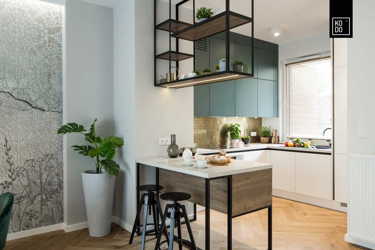 Nowoczesna kuchnia z wyspą, zielonymi szafkami i złotą mozaiką na ścianie, uzupełniona o czarne metalowe półki wiszące i dekoracyjną tapetę z motywami roślinnymi, oświetlona ciepłym światłem.