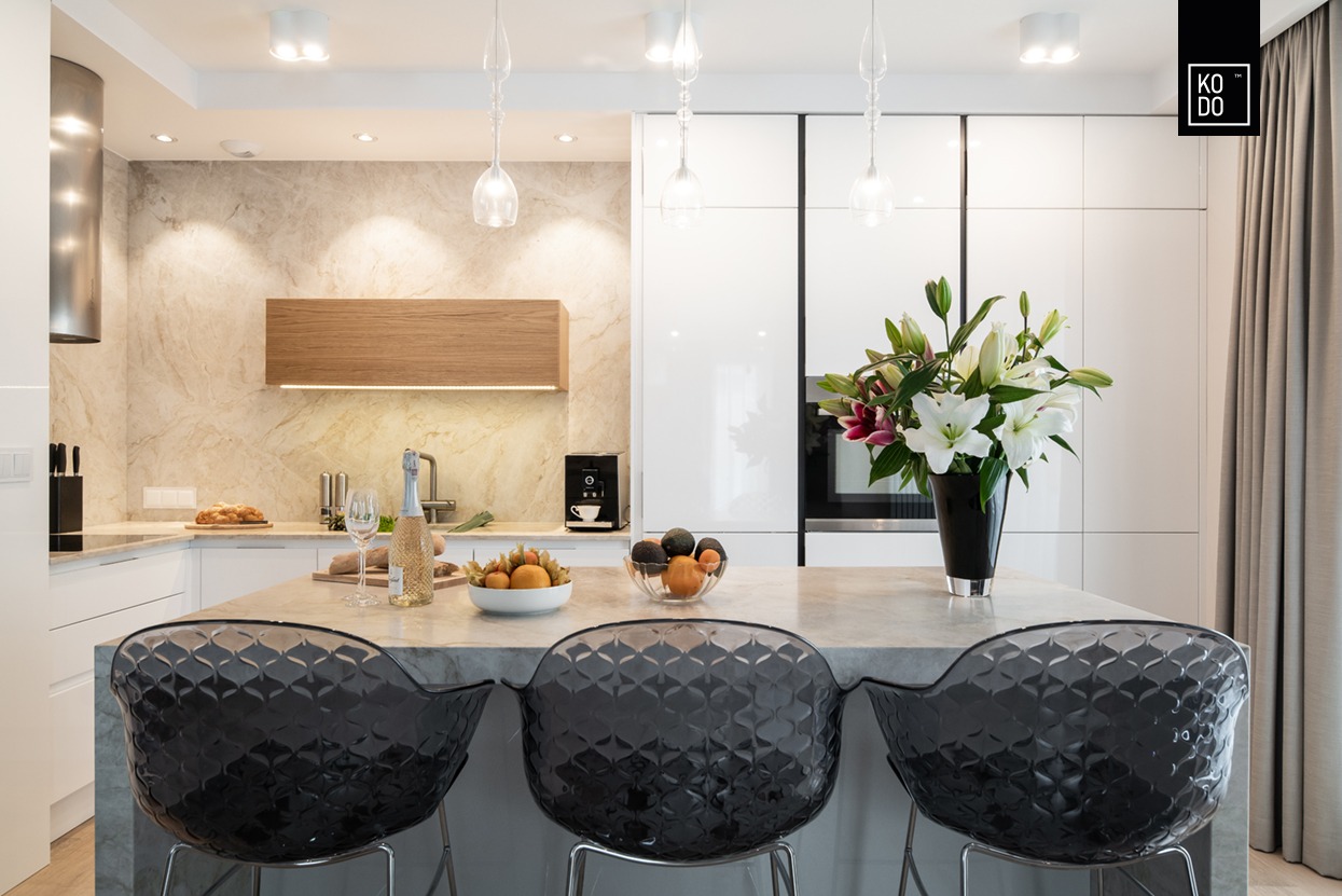 Nowoczesna kuchnia z wyspą, marmurowym blatem, transparentnymi krzesłami barowymi i bukietem lilii w czarnym wazonie, oświetlona wiszącymi lampami.