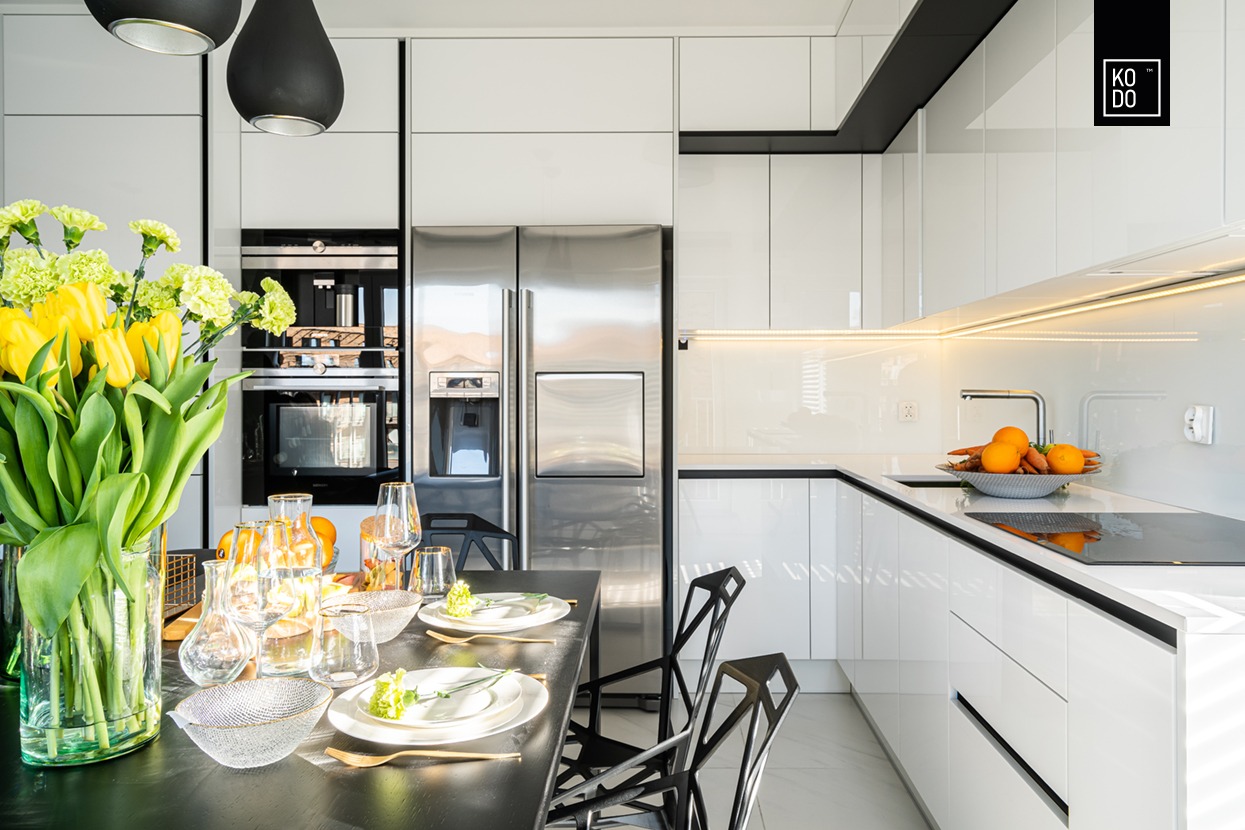 Nowoczesna, biała kuchnia z czarnym stołem jadalnym, na którym stoją kwiaty, szklanki i talerze. Widoczny srebrny sprzęt AGD i oświetlenie LED pod szafkami. W prawym górnym rogu logo firmy KODO.