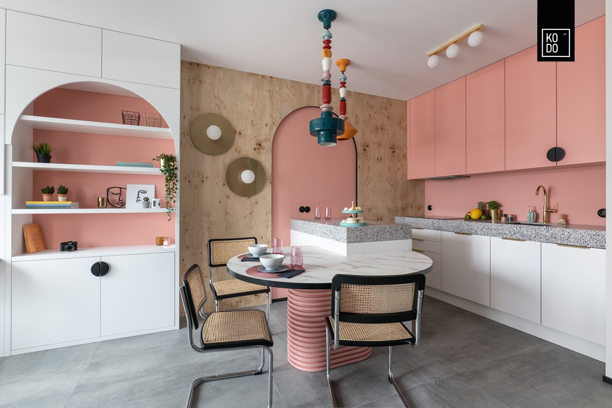 Nowoczesna kuchnia z różowymi szafkami, drewnianą ścianą, okrągłym stołem z marmurowym blatem i wiklinowymi krzesłami, oświetlona designerską lampą wiszącą i złotymi lampami ściennymi.