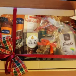 Kartonowe pudełko prezentowe z okienkiem, wypełnione słodyczami, kawą i przetworami, przewiązane czerwoną wstążką z ozdobną kokardą w kratę i drewnianymi gwiazdkami.