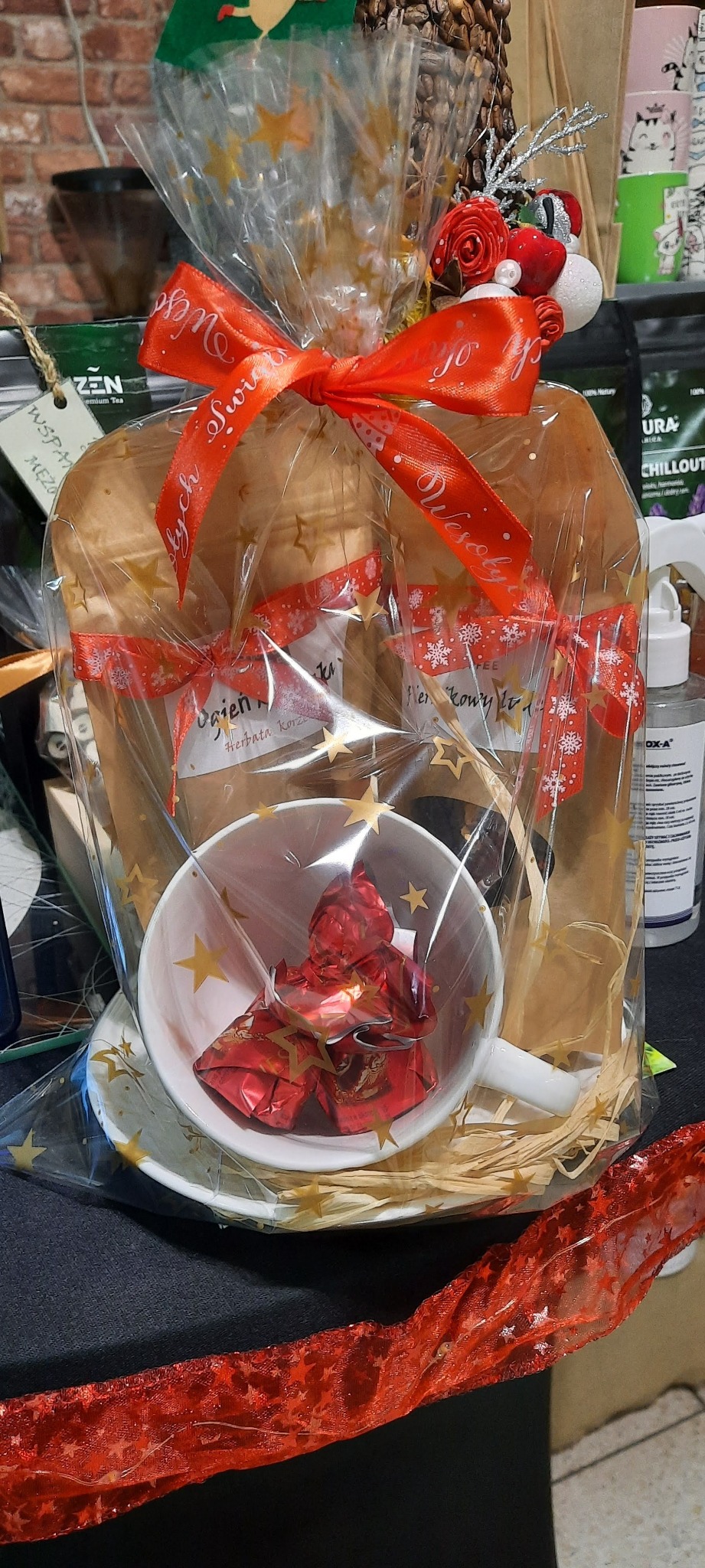 Udekorowany zestaw prezentowy z herbatą, kawą i słodyczami w filiżance, zapakowany w celofan z czerwoną kokardą z napisem 'Wesołych Świąt'.