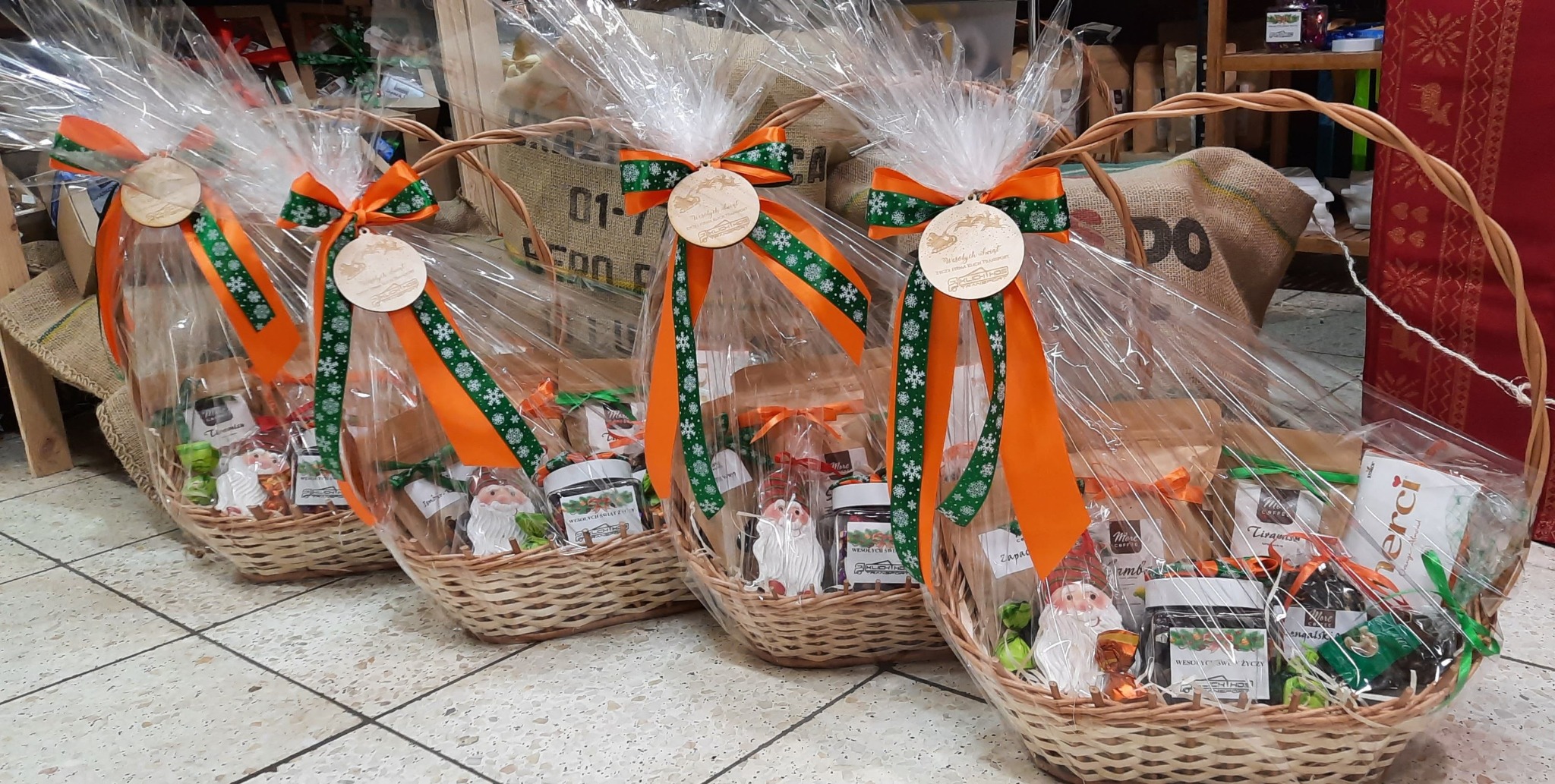 Trzy wiklinowe kosze zapakowane w folię, ozdobione pomarańczowymi wstążkami z motywem śnieżynek, drewnianymi zawieszkami z logo firmy oraz zawierające słodycze, kawy i figurki Mikołajów.
