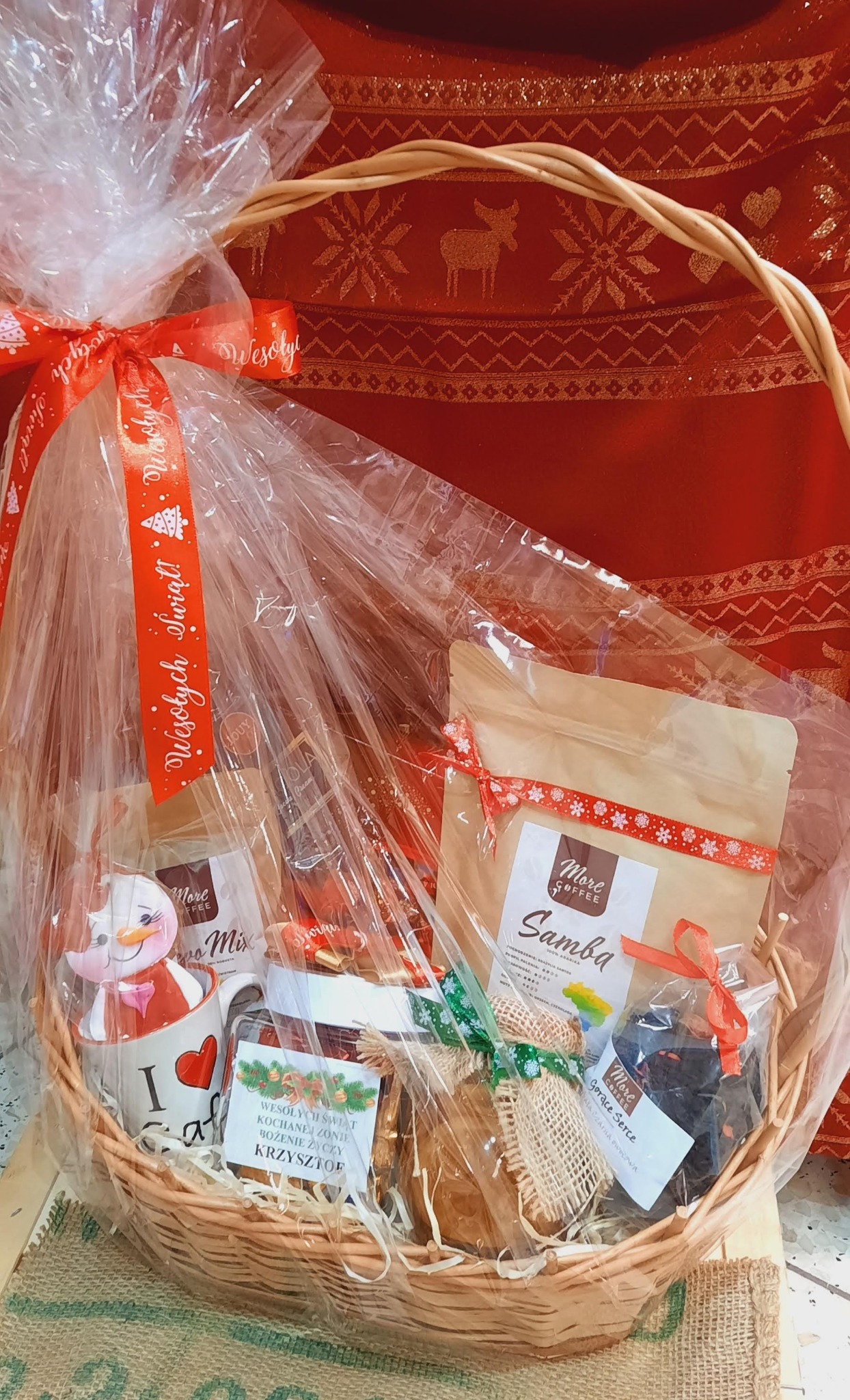 Wiklinowy kosz wypełniony artykułami spożywczymi i upominkami, zapakowany w przezroczystą folię i ozdobiony czerwoną wstążką z napisem 'Wesołych Świąt', na czerwonym tle z motywem reniferów...
