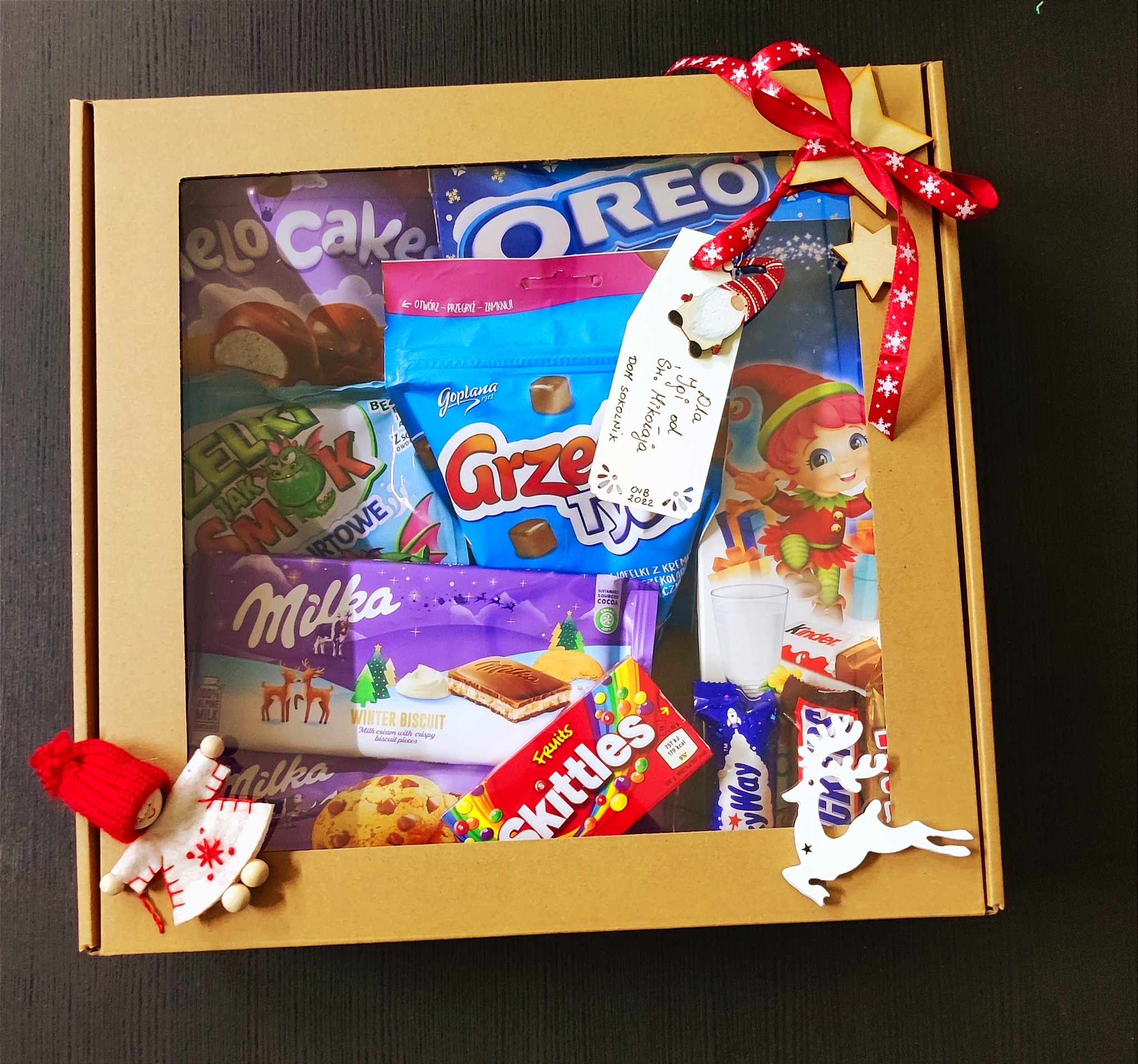 Kartonowe pudełko z okienkiem wypełnione różnorodnymi słodyczami, ozdobione czerwoną wstążką w białe śnieżynki i drewnianymi gwiazdkami, dodatkowo przyozdobione ręcznie robioną figurką aniołka...
