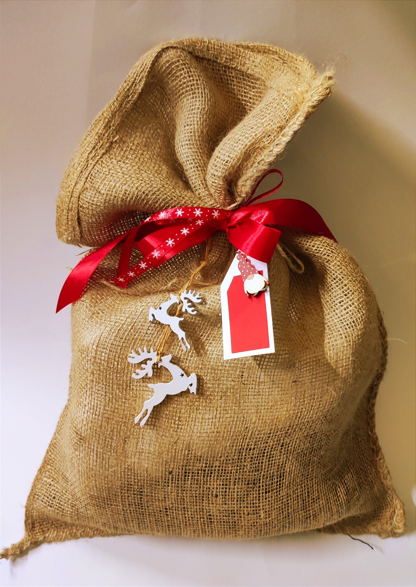 Jutowy worek prezentowy, przewiązany czerwoną wstążką w białe śnieżynki i ozdobiony zawieszkami w kształcie reniferów oraz czerwoną etykietą z dekoracyjnym krasnalem.