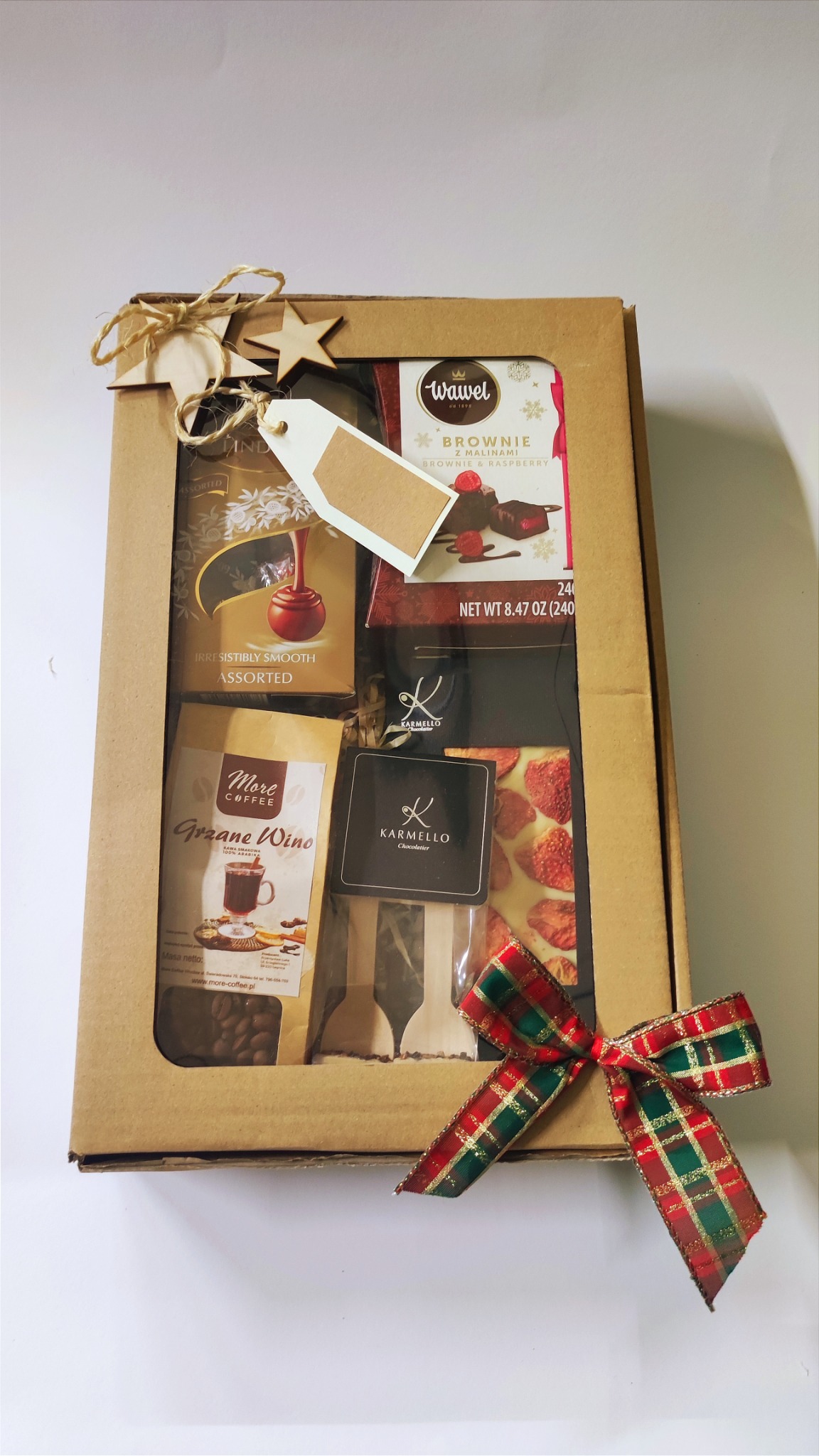 Kartonowe pudełko z okienkiem wypełnione kawą, czekoladą i dodatkami, udekorowane drewnianymi gwiazdkami, sznurkiem i świąteczną kokardą w kratę.