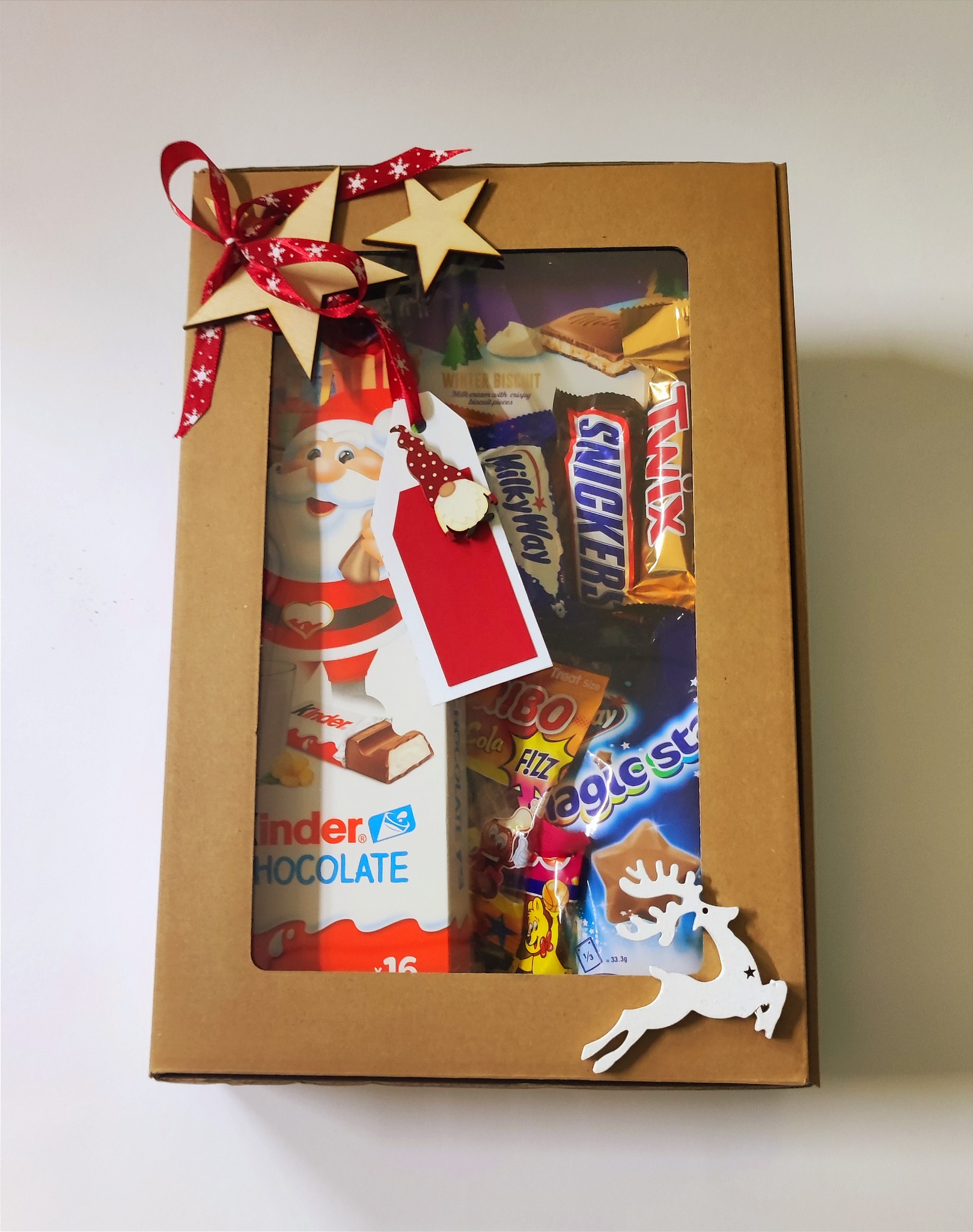 Kartonowe pudełko z okienkiem wypełnione słodyczami: Kinder Chocolate, Snickers, Twix, Milky Way, Jojo FIZZ Cola i Magic Stars, ozdobione drewnianymi gwiazdkami, czerwoną wstążką w białe śnieżynki,...