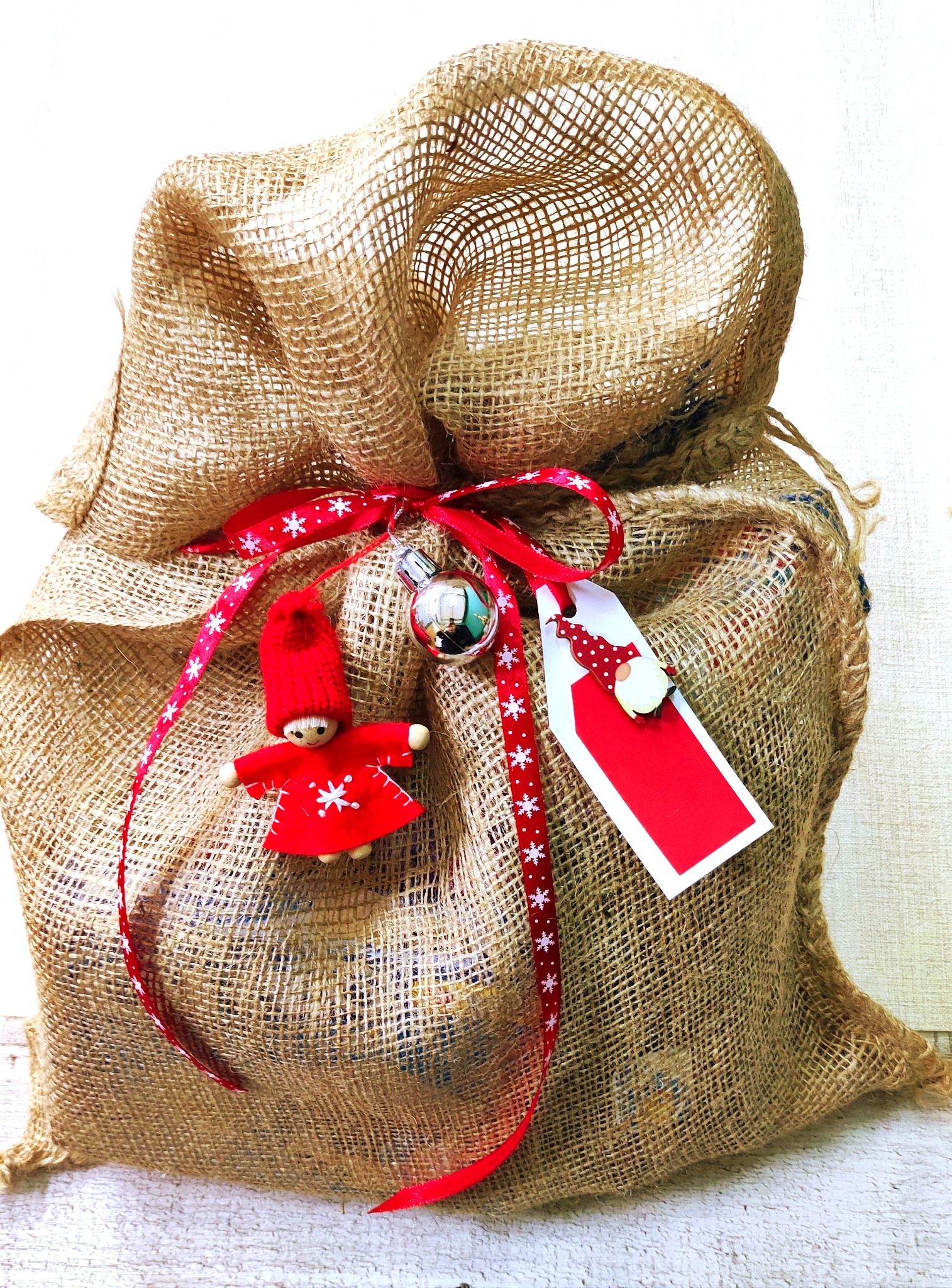 Jutowy worek zawiązany czerwoną wstążką w białe śnieżynki, z dołączoną srebrną bombką, czerwoną etykietą i małą, drewnianą laleczką w czerwonym stroju i czapce.